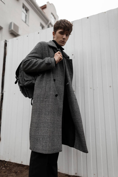 Elegant handsome young man in a fashionable plaid coat with a stylish leather backpack with a trendy hairstyle stands on the street near a modern metal white wall. Modern men's seasonal outerwear. - Zdjęcie, obraz