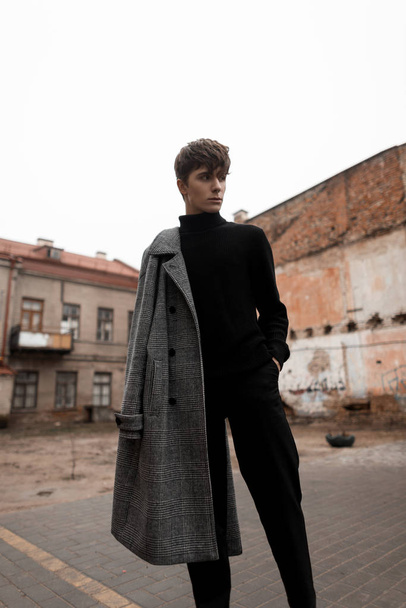 Jonge Europese man model met een modieuze kapsel in een stijlvolle grijze geruite jas in jeans in een zwarte vintage trui staat buiten op straat tussen de gebouwen. Leuke trendy europese kerel. - Foto, afbeelding