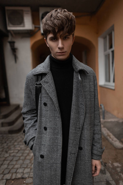 Νεαρός άνδρας μοντέλο σε ένα κομψό γκρι καρό παλτό σε μαύρο πλεκτό πουλόβερ με μοντέρνο χτένισμα ποζάρουν στην πόλη στην αυλή ενός κτιρίου εποχής. Ωραίος τύπος με κομψά ρούχα έξω. Στυλ. - Φωτογραφία, εικόνα