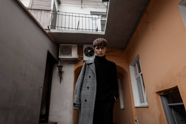 Elegante modieuze jongeman met een trendy kapsel in een zwarte vintage trui in een stijlvolle grijze geruite herfstjas staat in de stad vlakbij het gebouw. Knappe jongen model. Seizoensgebonden. - Foto, afbeelding