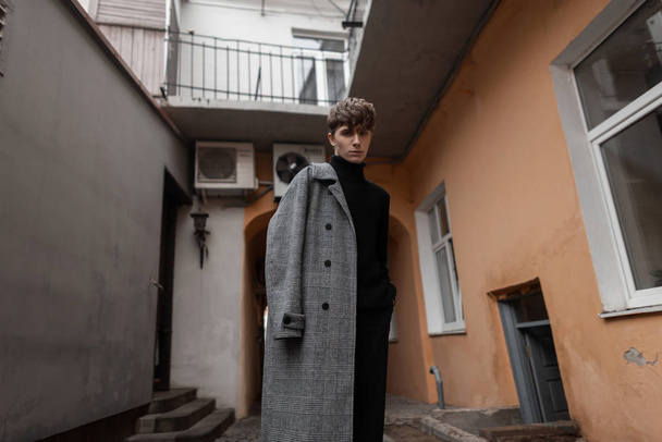Mladý módní sexy muž se stylovým účesem v klasickém šedém kostkovaném kabátě v módním černém pleteném svetru stojí na nádvoří vinobraní budovy. Módní model atraktivní městský chlap. - Fotografie, Obrázek