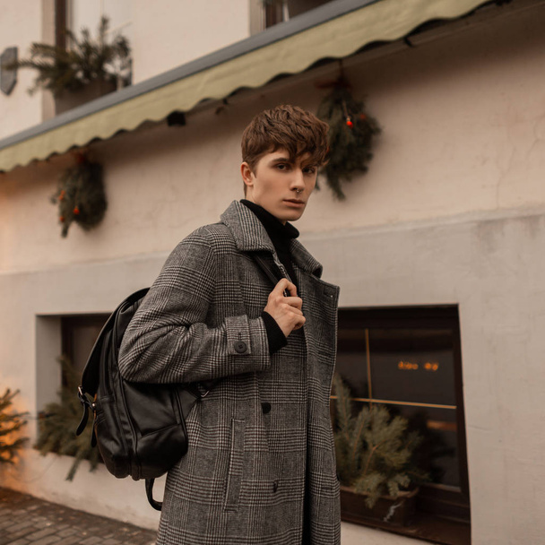 stilvoller junger Mann in schickem kariertem grauen Mantel mit modischem Lederrucksack spaziert die Straße entlang in der Nähe eines schönen Gebäudes mit Weihnachtsbaum und Vintage-Girlanden. - Foto, Bild