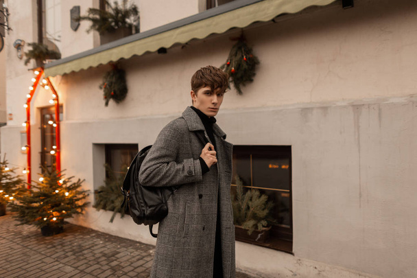 Divatos fiatalember divatos kockás kabátban, elegáns bőrtáskával sétál végig a város utcáján, közel egy karácsonyi koszorúkkal és fákkal díszített épülethez. Jóképű srác élvezi a sétát.. - Fotó, kép