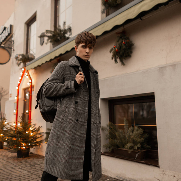 European young man in a trendy plaid coat with a stylish leather backpack walks along a city street near a building decorated with Christmas garlands and trees. Modern handsome guy is enjoy a walk. - Photo, image