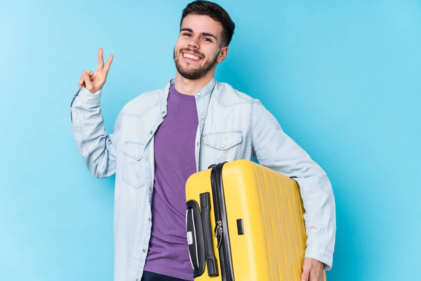 Joven viajero caucásico sosteniendo una maleta aislada alegre y despreocupada mostrando un símbolo de paz con los dedos
. - Foto, imagen
