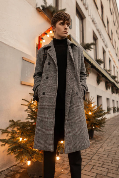Bonito jovem bonito em um elegante casaco xadrez cinza está de pé perto do edifício entre duas belas árvores de Natal. Europeu cara modelo goza de um passeio na cidade no fim de semana de Ano Novo
. - Foto, Imagem