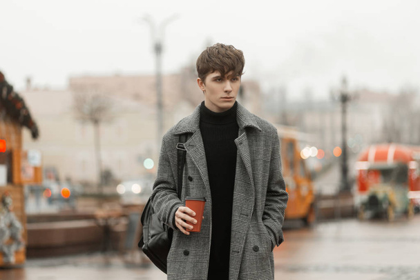 Модель модного молодого человека в элегантном молодежном клетчатом пальто с модной прической стоит на городской площади с горячим напитком в руках. Городской парень выходит на улицу и пьет сладкий чай.
. - Фото, изображение