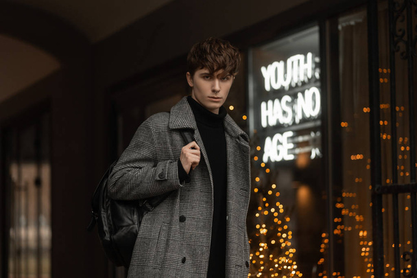 Young man with a trendy hairstyle in a gray checkered seasonal fashionable coat with a stylish backpack on shoulder is stands on the street near a shop window with bright lights. Guy model in the city - Photo, Image