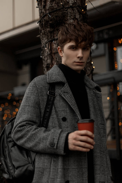 Joven guapo en un elegante abrigo a cuadros con una mochila de moda se levanta con una taza de delicioso café en la calle. Atractivo chico pasea por la ciudad en un fin de semana festivo con una bebida caliente
. - Foto, Imagen