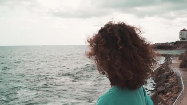 Όμορφη σγουρή μελαχρινή γυναίκα κοιτάζει τη θάλασσα σε μια συννεφιασμένη μέρα. - Πλάνα, βίντεο