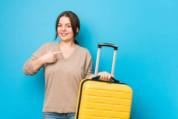 Young curvy woman holding a suitcase smiling and pointing aside, showing something at blank space. - Photo, Image