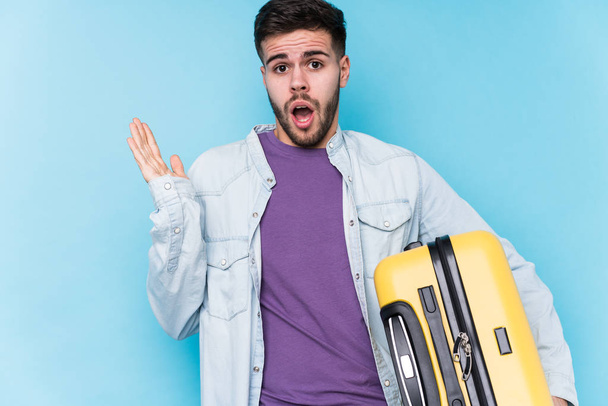 Νεαρός καυκάσιος ταξιδιώτης κρατώντας μια βαλίτσα απομονωμένη έκπληκτος και σοκαρισμένος. - Φωτογραφία, εικόνα