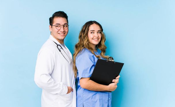 Joven pareja de médicos posando en un fondo azul aislado se ve a un lado sonriente, alegre y agradable
. - Foto, Imagen