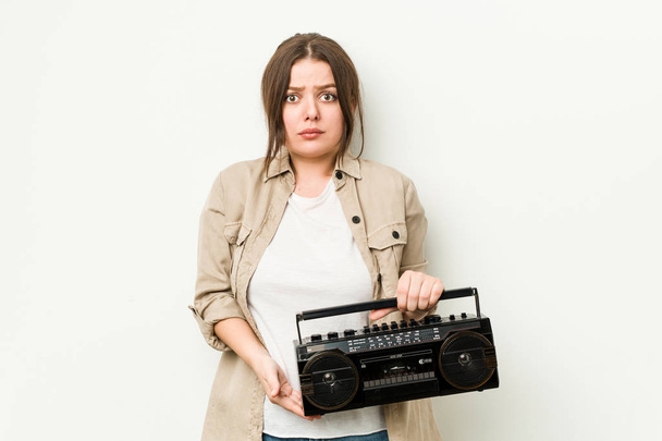 Young curvy woman holding a retro radio shrugs shoulders and open eyes confused. - Photo, Image