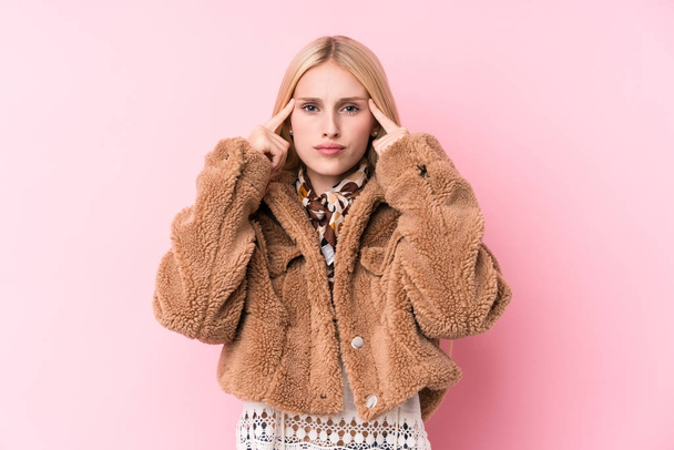 Young blonde woman wearing a coat against a pink background focused on a task, keeping forefingers pointing head. - 写真・画像