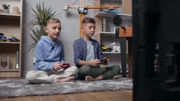 Knappe geconcentreerde tiener jongens zitten op het tapijt en spelen video game met behulp van joysticks - Video
