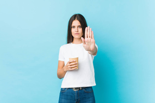 Jonge blanke vrouw met een afhaalkoffie staande met uitgestrekte hand met stopteken, voorkomen dat u. - Foto, afbeelding