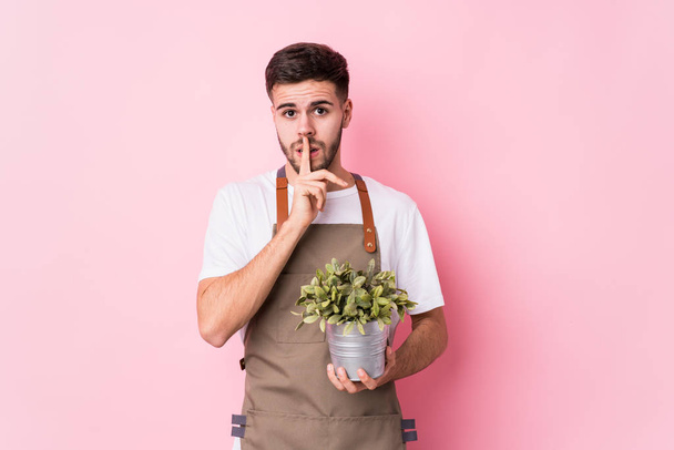 Young caucasian gardener man holding a plant isolated keeping a secret or asking for silence. - Photo, Image