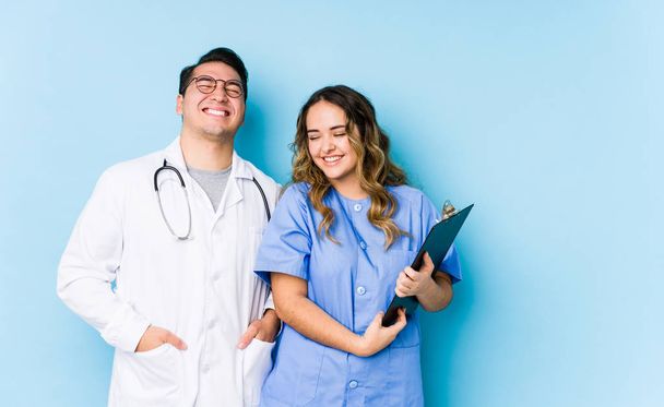 junges Ärztepaar posiert isoliert vor blauem Hintergrund, lacht und schließt die Augen, fühlt sich entspannt und glücklich. - Foto, Bild
