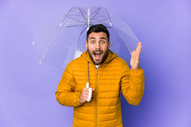 Młody przystojny mężczyzna trzymając parasol odizolowany otrzymuje miłą niespodziankę, podekscytowany i podnoszące ręce. - Zdjęcie, obraz