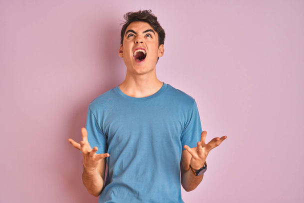 Tiener jongen draagt casual t-shirt staan over blauw geïsoleerde achtergrond gek en gek schreeuwen en schreeuwen met agressieve uitdrukking en armen omhoog. Frustratieconcept. - Foto, afbeelding