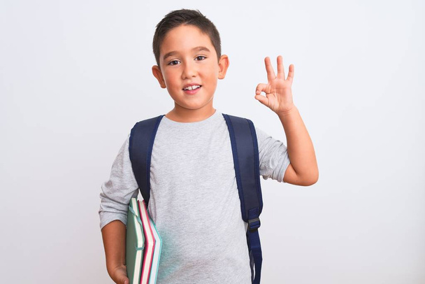 Piękny uczeń chłopiec nosi plecak trzymając książki na odizolowanym białym tle robi ok znak palcami, doskonały symbol - Zdjęcie, obraz