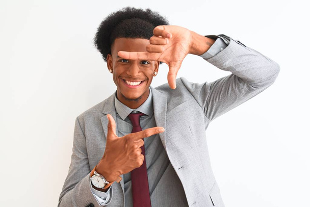 Mladý africký americký podnikatel v obleku stojí nad osamělým bílým pozadím a usmívá se a vytváří rámeček s rukama a prsty se šťastnou tváří. Koncept kreativity a fotografie. - Fotografie, Obrázek