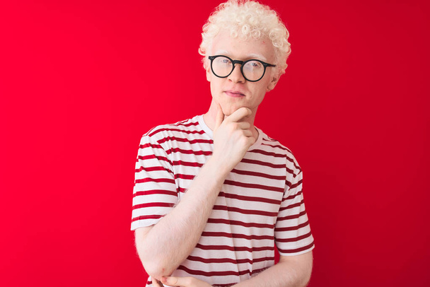 Jonge albino blonde man met gestreept t-shirt en bril over geïsoleerde rode achtergrond, zelfverzekerd kijkend naar de camera met glimlach met gekruiste armen en hand geheven op kin. Positief denken. - Foto, afbeelding