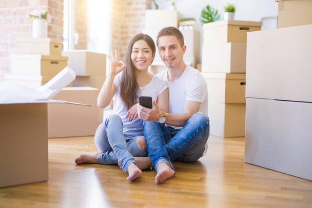 Beau couple assis sur le sol en utilisant un smartphone à la nouvelle maison autour de boîtes en carton faisant signe ok avec les doigts, excellent symbole
 - Photo, image