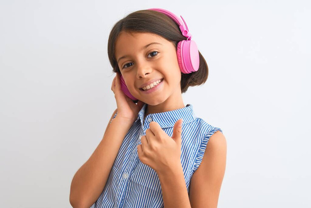 Gyönyörű gyermek lány zenét hallgat fülhallgató segítségével elszigetelt fehér háttér boldog nagy mosollyal csinál oké jel, hüvelykujj fel ujjaival, kiváló jel - Fotó, kép