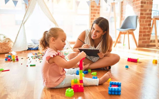 Beautiful psycologist and blond toddler girl doing therapy building tower using plastic blocks at kindergarten - Φωτογραφία, εικόνα
