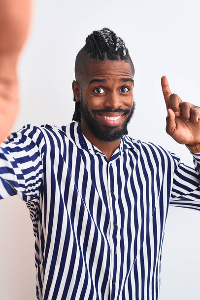 Hombre afroamericano con trenzas hacen selfie por cámara sobre fondo blanco aislado sorprendido con una idea o pregunta señalando el dedo con la cara feliz, número uno
 - Foto, imagen