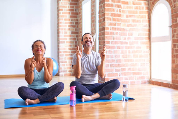 Sportliches Paar mittleren Alters, das auf einer Matte sitzt und Stretching Yoga-Übungen im Fitnessstudio macht und mit gekreuzten Fingern lächelnd und geschlossenen Augen macht. Glück und abergläubisches Konzept. - Foto, Bild