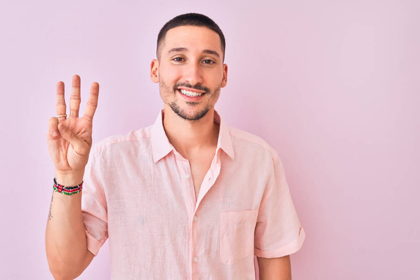 Homem bonito jovem vestindo camisa rosa em pé sobre fundo isolado mostrando e apontando para cima com os dedos número três, sorrindo confiante e feliz
. - Foto, Imagem