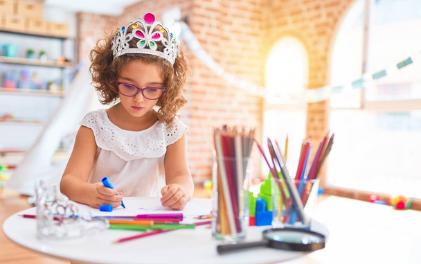 Beautiful toddler wearing glasses and princess crown sitting drawing using paper and marker pen at kindergarten - Foto, Imagem