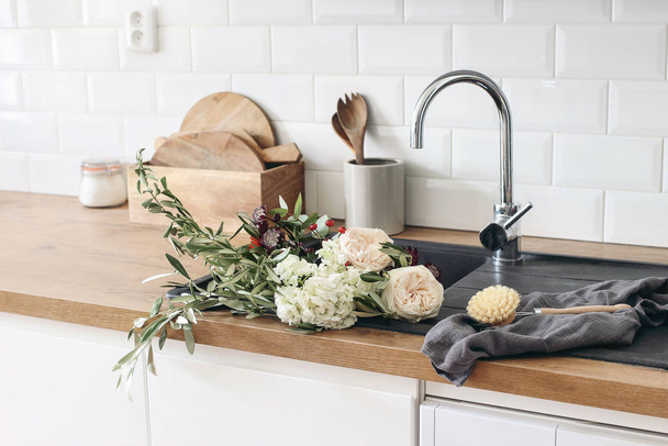 Closeup of kitchen interior. White brick wall, metro tiles, wooden countertops with kitchen utensils. Roses flowers in black sink. Modern scandinavian design. Home staging, cleaning concept. - Photo, image