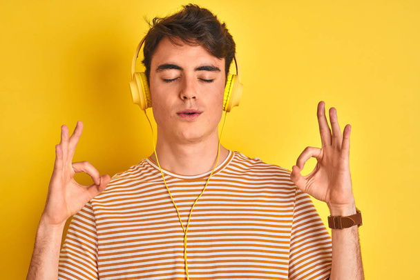 Teenager chlapec nosí sluchátka přes izolované žluté pozadí relaxovat a usmívat se se zavřenýma očima dělat meditační gesto s prsty. Koncept jógy. - Fotografie, Obrázek