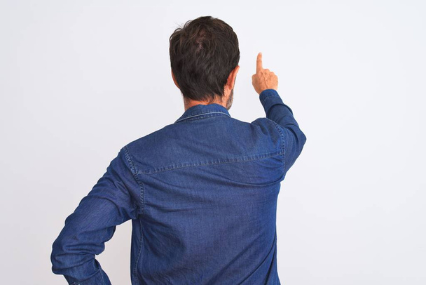 Мужчина средних лет в синей джинсовой рубашке стоял на изолированном белом фоне, задом наперёд показывая пальцем вперед.
 - Фото, изображение