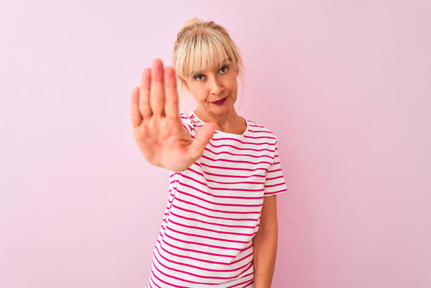 Donna di mezza età che indossa t-shirt a righe in piedi su sfondo rosa isolato facendo smettere di cantare con il palmo della mano. Espressione di avvertimento con gesto negativo e serio sul viso
. - Foto, immagini