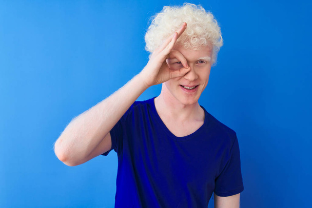 Young albino blond man wearing casual t-shirt standing over isolated blue background doing ok gesture with hand smiling, eye looking through fingers with happy face. - Фото, изображение