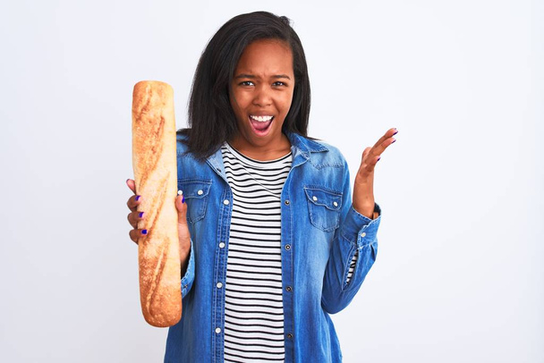 Joven mujer afroamericana sosteniendo pan sobre un fondo aislado muy feliz y emocionada, expresión ganadora celebrando la victoria gritando con una gran sonrisa y levantando las manos
 - Foto, imagen