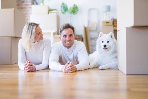 Young beautiful couple with dog lying down on the floor at new home around cardboard boxes - Photo, Image