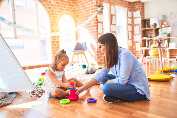 白人の女の子の子供は女教師と遊び場で遊んで学びます.Mother and daughter at playroom playing with inteligence toys - 写真・画像