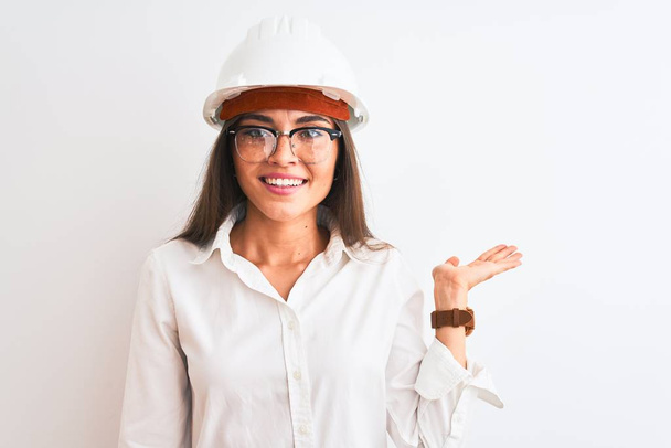 Молодая красивая архитектор женщина в шлеме и в очках на изолированном белом фоне улыбается веселым подарком и указывает пальцем на камеру
. - Фото, изображение