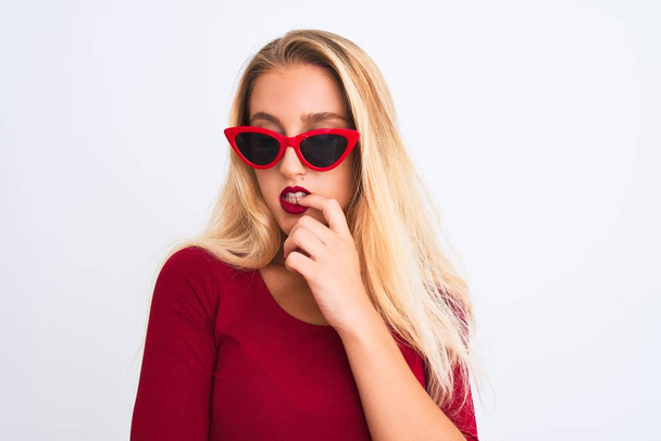 Young beautiful woman wearing red t-shirt and sunglasses over isolated white background looking stressed and nervous with hands on mouth biting nails. Anxiety problem. - Photo, image