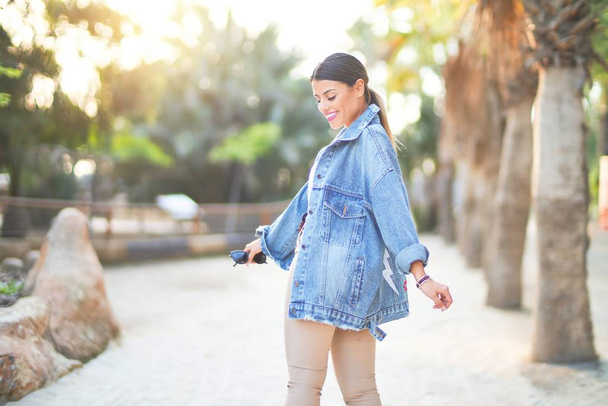 Молодая красивая женщина в джинсовой куртке улыбается счастливой и уверенной. Стоя с улыбкой на лице в городском парке
 - Фото, изображение