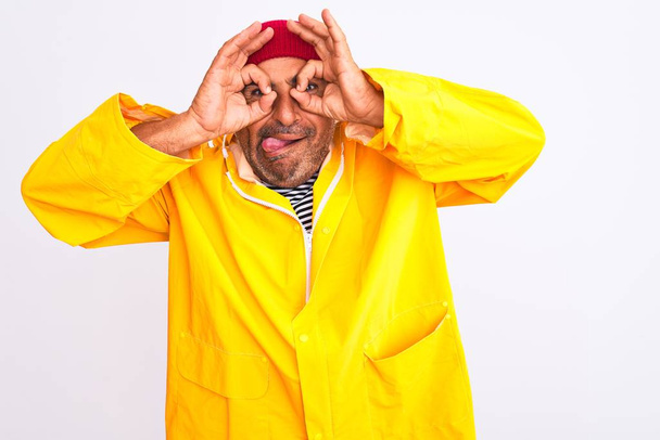 Middle age man wearing rain coat and woolen hat standing over isolated white background doing ok gesture like binoculars sticking tongue out, eyes looking through fingers. Crazy expression. - Foto, Bild