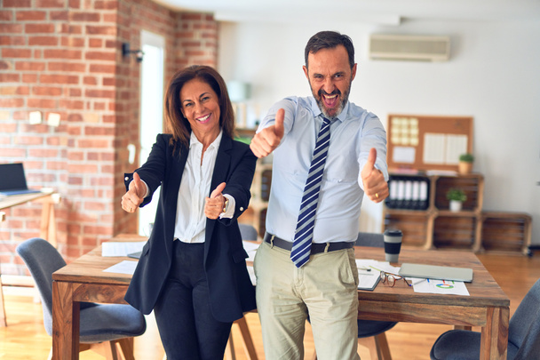 Dwóch pracowników średniego wieku pracujących razem na spotkaniu w biurze, zatwierdzających pozytywny gest ręką, uśmiechniętych i szczęśliwych z powodu sukcesu. Gest zwycięzcy. - Zdjęcie, obraz