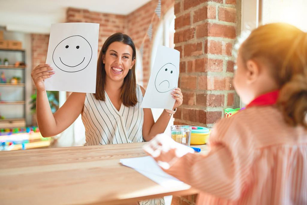 Beautiful psycologist and blond toddler girl wearing school uniform sitting on desk doing therapy using emoji emotions at consulting room - Photo, Image