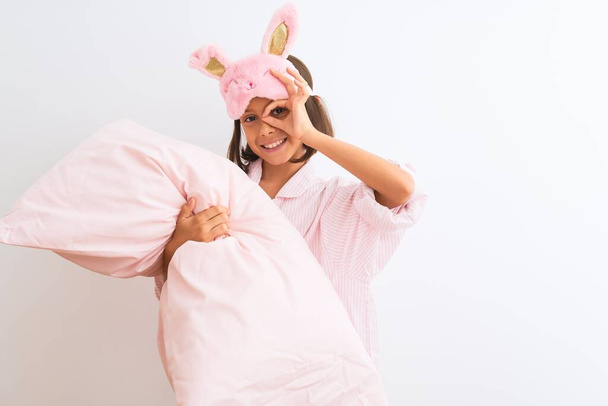 Kindermädchen mit Schlafmaske und Pyjama hält Kissen über isoliertem weißem Hintergrund mit glücklichem Gesicht lächelnd tut ok Zeichen mit Hand auf Auge durch Finger schauen - Foto, Bild